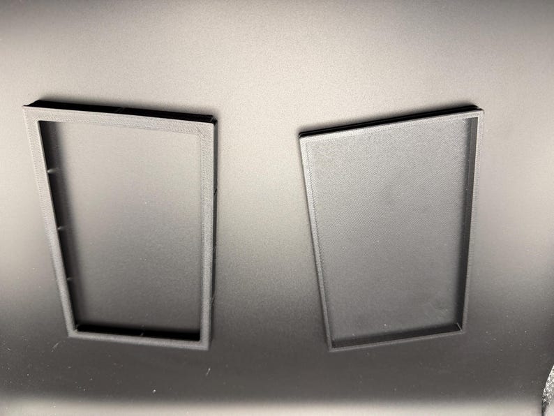 May include: Two black rectangular trays, one with an open frame and the other with a solid base, are displayed against a dark background. The trays appear to be made of a matte material, possibly plastic or a similar substance.