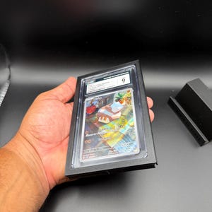 May include: A graded trading card encased in a black holder. The card features colorful artwork and Japanese text. The holder is marked with a "9" grade. The card is held in a person's hand against a dark background.
