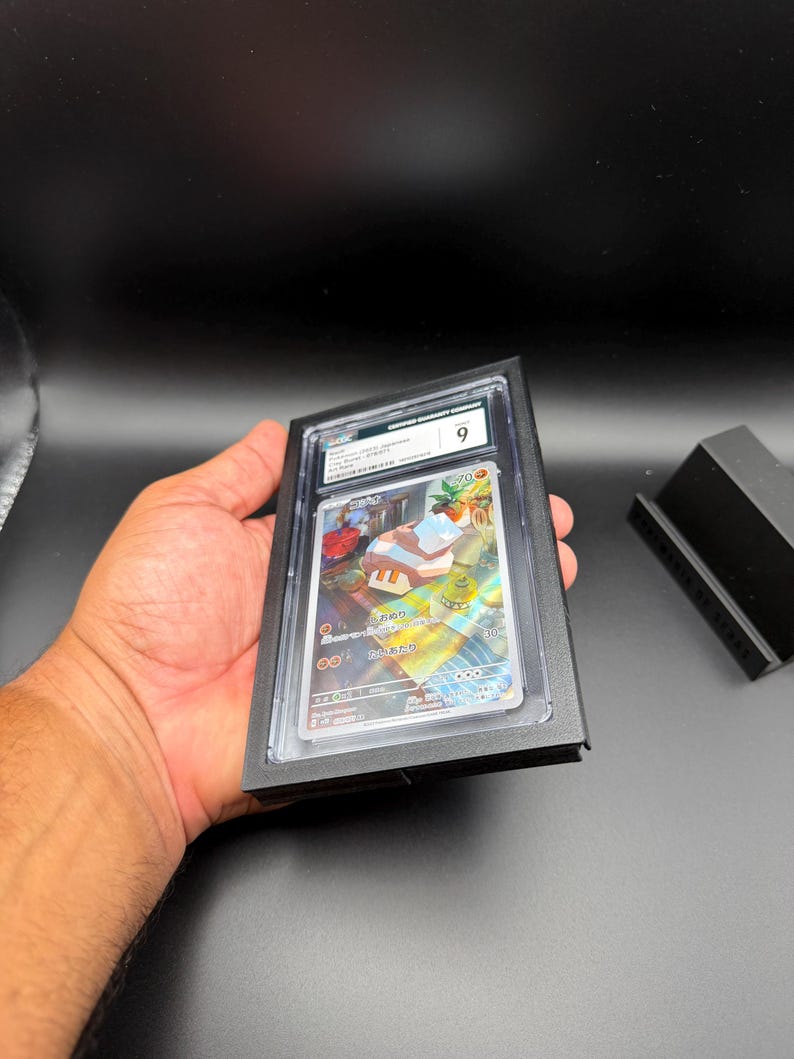May include: A graded trading card encased in a black holder. The card features colorful artwork and Japanese text. The holder is marked with a "9" grade. The card is held in a person's hand against a dark background.