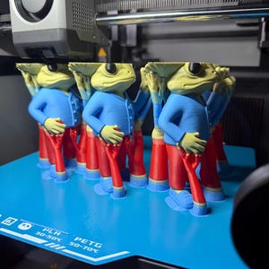 Puede incluir: Una impresora 3D está en funcionamiento, produciendo múltiples figuras de un personaje de rana. Las figuras visten chaquetas azules y pantalones rojos. La cabeza de la rana es de color verde claro y la base es azul.