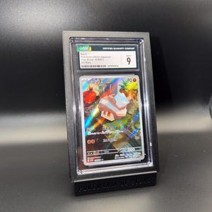 May include: A graded Pok&eacute;mon card in a black display case. The card features Japanese text and a colorful illustration. The case is labeled "CERTIFIED GUARANTY COMPANY" and has a grade of 9. The base of the display reads "POKEMANIA DE SEBAS."