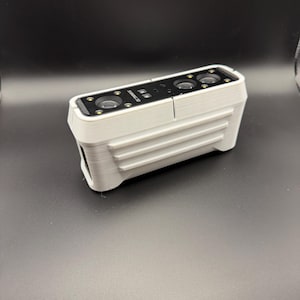 May include: A light gray 3D scanner with the text "SHINING 3D" printed on the top. The scanner has multiple lenses and a ridged design on the side. The device is on a dark gray surface.