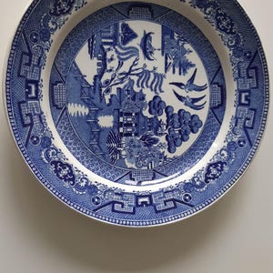 Op de afbeelding: Een ronde, wit en blauwe keramische plaat met een gedetailleerd Willow-patroon. De plaat heeft een centrale scène met een brug, vogels en een pagode, omgeven door een decoratieve rand. De plaat heeft een diameter van ongeveer 25 cm.