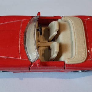 Op de afbeelding: Een felrode speelgoedcabriolet met een doorzichtige plastic voorruit, een zwart stuurwiel en twee beige stoelen. Het voertuig heeft een opgevouwen beige kapafdekking achter de stoelen en een gladde, glanzende afwerking aan de buitenkant.