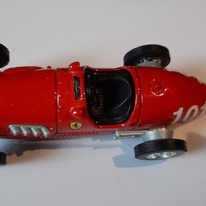 Op de afbeelding: Een rode speelgoedraceauto van metaal met het nummer 101 in het wit op de neus, zwarte banden, zilveren wieldoppen en een klein Ferrari-embleem aan de zijkant. De open cockpit toont een zwart interieur en zilverkleurige uitlaatpijpen.