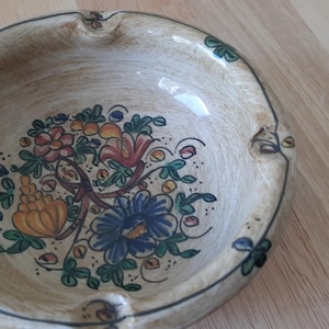 Op de afbeelding: Een beige keramische asbak met een bloemenontwerp. De asbak heeft een ronde vorm met drie inkepingen om sigaretten op te leggen. Het bloemenontwerp toont blauwe, oranje en rode bloemen met groene bladeren.