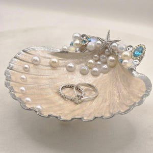 Puede incluir: Un soporte decorativo para anillos con forma de concha marina y borde plateado. El interior está lleno de perlas de imitación, una estrella de mar y dos anillos plateados. Incluye cristales azules y transparentes.
