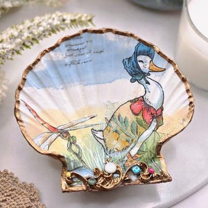 Op de afbeelding: Een decoratieve schelpschaal met een illustratie van een eend met een muts en jurk, en een libelle. De schelp heeft een gouden rand en is versierd met kleine, kleurrijke versieringen.