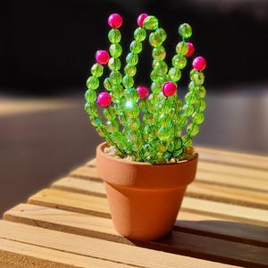 May include: A beaded cactus ornament in a terracotta pot. The cactus is made of green beads with pink accents. The pot is placed on a wooden surface, with a blurred background.