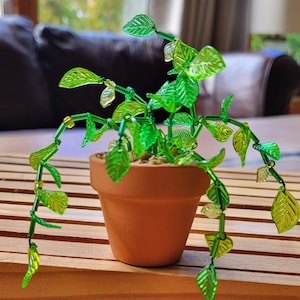 May include: A terracotta plant pot containing a decorative plant with green and yellow glass leaves. The plant's stems and leaves are made of glass beads, creating a unique, handcrafted look. The pot sits on a wooden surface.