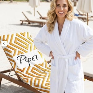 May include: A white terry cloth robe with a matching belt and pockets. A beach chair with a white and gold geometric patterned towel that has the name "Piper" printed on it. The background shows a beach scene with lounge chairs and umbrellas.