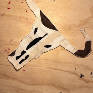 Può includere: Un appendiabiti decorativo a forma di teschio di mucca. L'appendiabiti presenta un tessuto color crema con dettagli marroni e neri. Il design ricorda un'estetica rustica e occidentale.