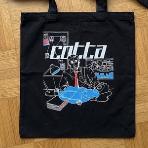 Op de afbeelding: Een zwarte draagtas met een witte grafische print van een persoon die aan een bureau zit met een telefoon, een lamp en documenten. De tekst "cotta" staat in grote witte letters boven de figuur. Het ontwerp bevat blauwe en rode accenten.