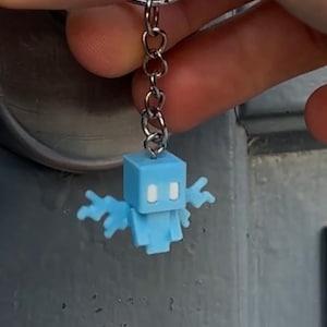 May include: A light blue keychain featuring a pixelated character with outstretched arms. The character is attached to a silver chain and keyring. The keychain is held up against a grey background.