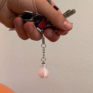 Könnte beinhalten: Eine Sammlung von Schlüsseln, die mit einem silbernen Schlüsselring zusammengehalten werden. Ein kleiner, rosa, dekorativer Flaschenanhänger hängt am Schlüsselring. Der Anhänger hat ein strukturiertes, geometrisches Design.
