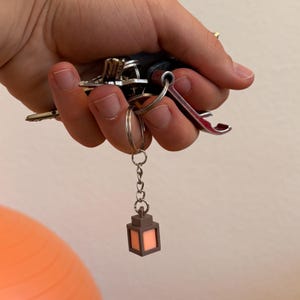 Puede incluir: Un llavero con un conjunto de llaves, que incluye una llave de coche y un abrebotellas. Un pequeño dije de linterna marrón con un brillo naranja cuelga del llavero. Las llaves se sostienen en una mano.