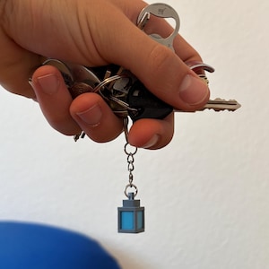 Peut inclure: Une main tenant un trousseau de clés, dont une clé de voiture et un ouvre-bouteille. Un petit porte-clés en forme de lanterne grise avec un intérieur bleu est attaché aux clés.