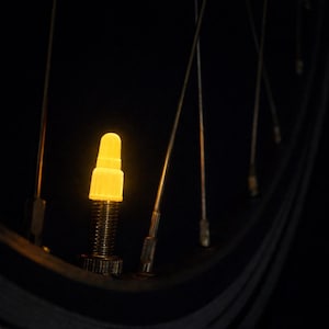 Puede incluir: Primer plano de un tapón de válvula de neumático de bicicleta amarillo brillante. El tapón está iluminado, contrastando con el fondo oscuro. Los radios de la rueda son visibles, agregando detalles.