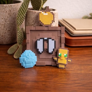 Könnte beinhalten: Eine Sammlung pixeliger Minecraft-Figuren. Enthält eine braune Truhe, einen blauen Würfel, einen gelben Apfel und eine kleine Figur mit gelbem Kopf und grünen Augen. Auf einer Holzoberfläche angeordnet.
