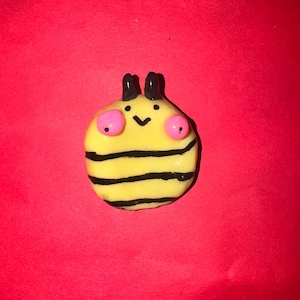 May include: A whimsical, handmade bee-shaped ornament. The ornament features a yellow body with black stripes, pink eyes, and black antennae. The background is a solid red color.