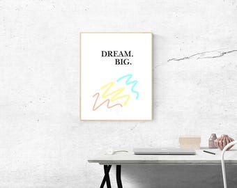 May include: A rectangular wall art print in a light wood frame featuring the black text DREAM. BIG. above three abstract, hand-drawn scribbles in light pink, pale yellow, and bright cyan colours on a clean white background.