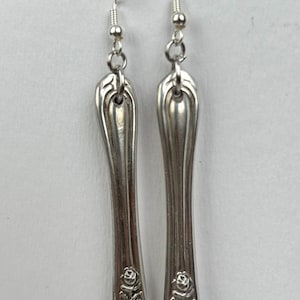 Puede incluir: Un par de pendientes de color plateado. Cada pendiente tiene forma de cuchara, con un diseño floral en la base. Los pendientes cuelgan de un gancho plateado y una pequeña cuenta.