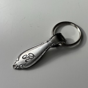 Op de afbeelding: Een zilverkleurige sleutelhanger in de vorm van een lepel met een gegraveerde letter "B" en bloemendetails. De sleutelhanger heeft een sleutelring bevestigd aan het handvat van de lepel. De lepel heeft een vintage design.