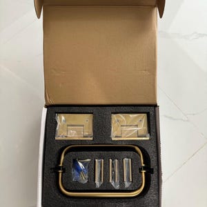 May include: A new, open cardboard box containing brass-colored shower door hardware. The set includes hinges, a handle, and mounting hardware, all nestled in a black foam insert. The hardware has a brushed finish.
