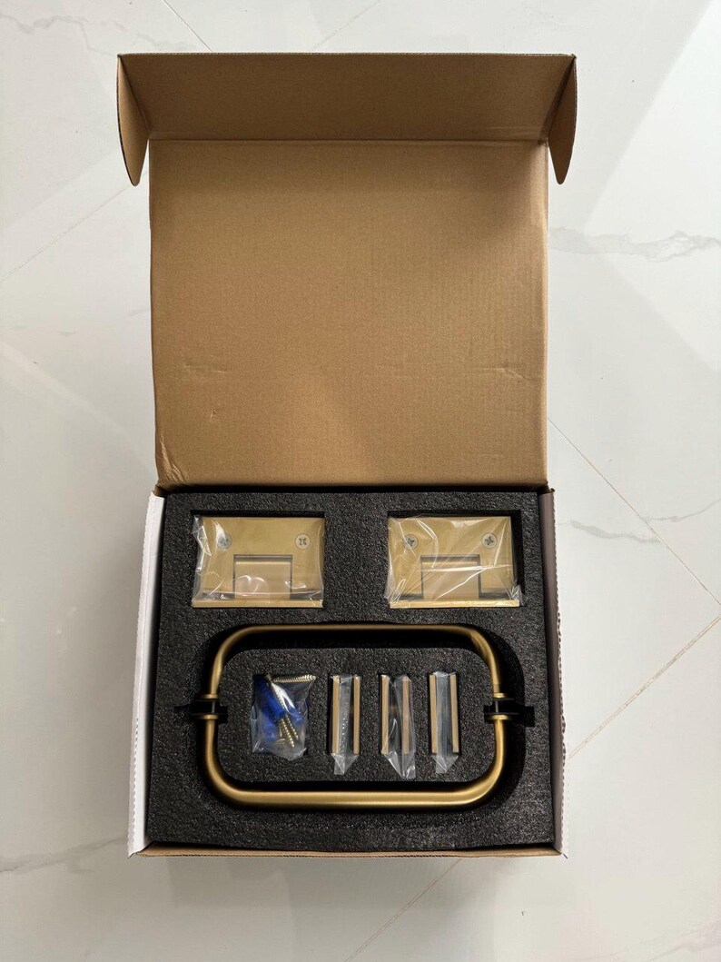 May include: A new, open cardboard box containing brass-colored shower door hardware. The set includes hinges, a handle, and mounting hardware, all nestled in a black foam insert. The hardware has a brushed finish.