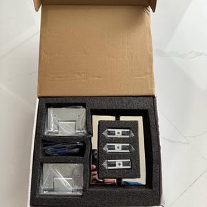 May include: An open cardboard box containing hardware for a glass door. The box includes two sets of hinges, three door clamps, and a small tool, all in a black foam insert. The hardware is made of shiny, silver-colored metal.