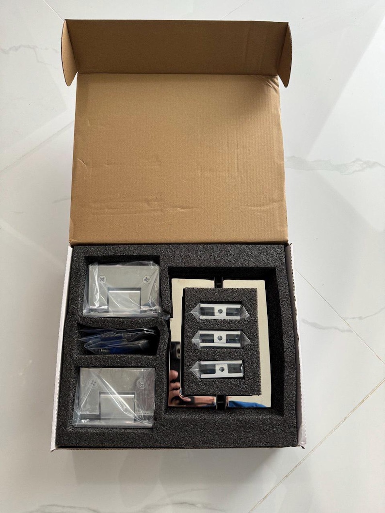 May include: An open cardboard box containing hardware for a glass door. The box includes two sets of hinges, three door clamps, and a small tool, all in a black foam insert. The hardware is made of shiny, silver-colored metal.