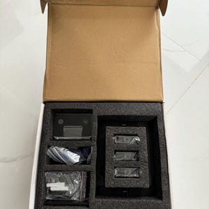 May include: An open cardboard box containing a black electronic device with a screen, three smaller black devices, and several small plastic bags. The items are nestled in a black foam insert.