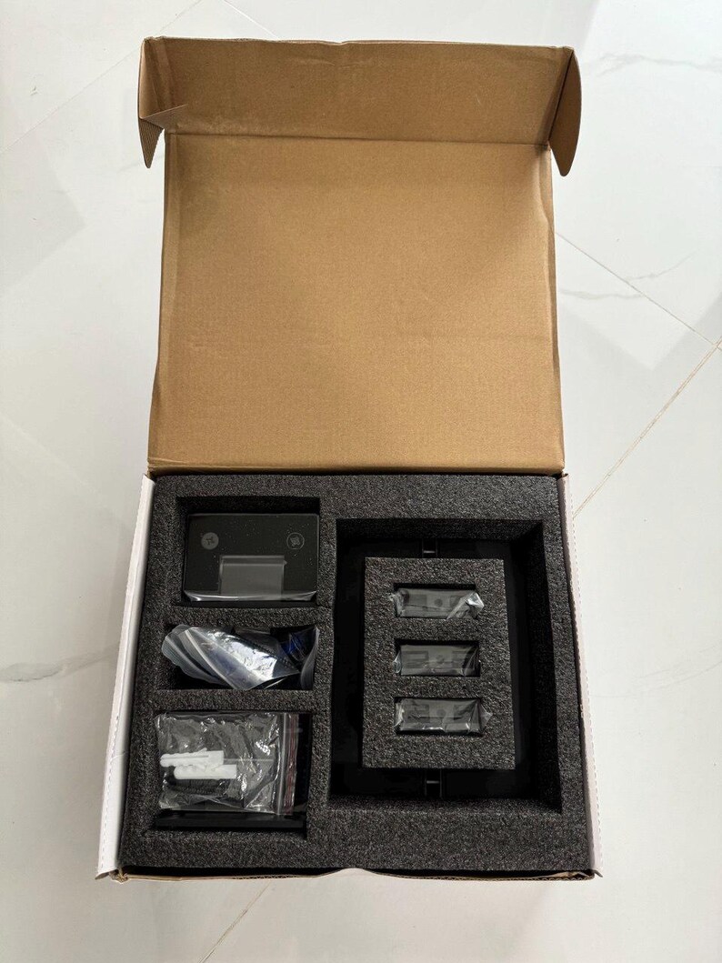 May include: An open cardboard box containing a black electronic device with a screen, three smaller black devices, and several small plastic bags. The items are nestled in a black foam insert.