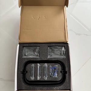 May include: An open cardboard box containing a black metal frame, several clear plastic-wrapped components, and a blue item. The items are nestled in a black foam insert.