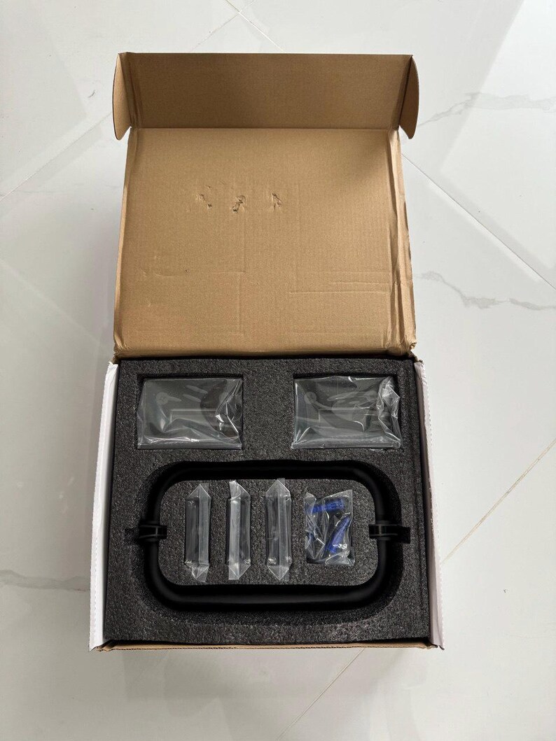 May include: An open cardboard box containing a black metal frame, several clear plastic-wrapped components, and a blue item. The items are nestled in a black foam insert.