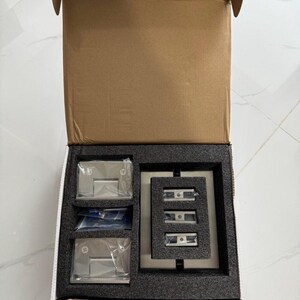 May include: An open cardboard box containing hardware components. Inside, a black foam insert holds several clear plastic-wrapped metal pieces, including hinges and mounting brackets. The box is on a white tiled surface.
