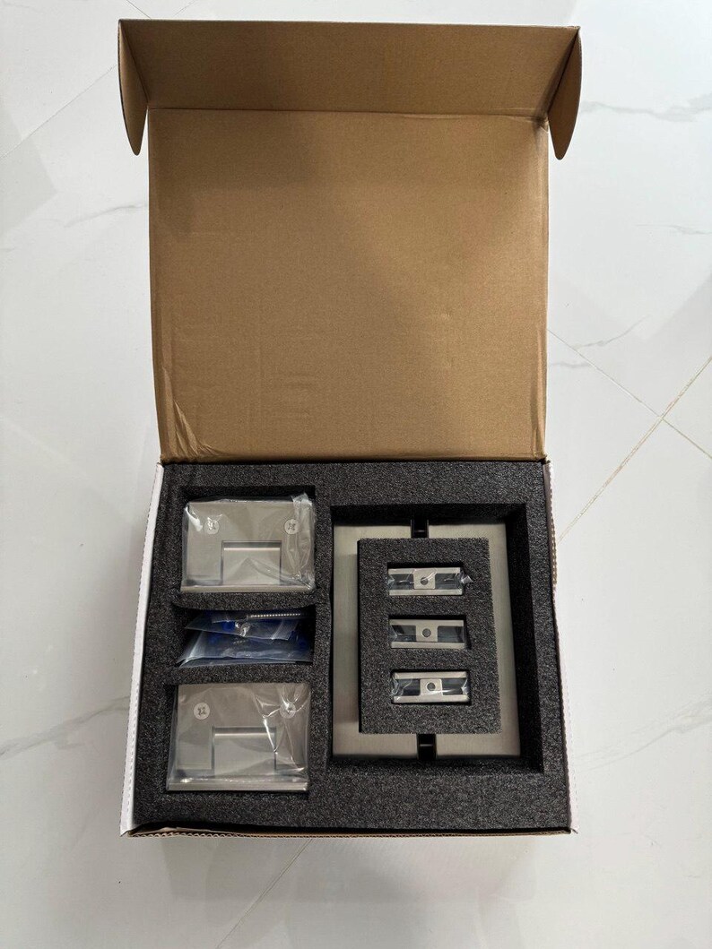 May include: An open cardboard box containing hardware components. Inside, a black foam insert holds several clear plastic-wrapped metal pieces, including hinges and mounting brackets. The box is on a white tiled surface.