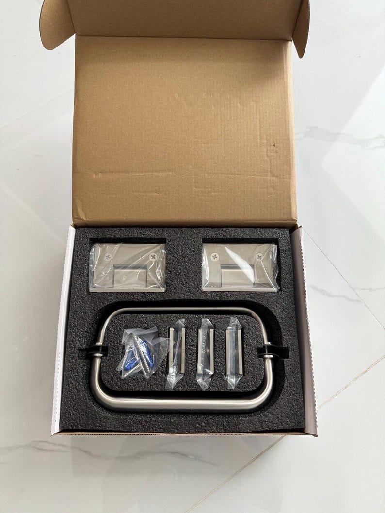 May include: A stainless steel shower door hardware kit, presented in an open cardboard box. The kit includes hinges, a handle, and mounting hardware, all neatly arranged in a black foam insert. The components are sealed in clear plastic.