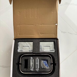 May include: An open cardboard box containing shower door hardware. The set includes two square, clear-wrapped hinges, a curved metal handle, and several small, clear, and blue-wrapped components, all nestled in a black foam insert.