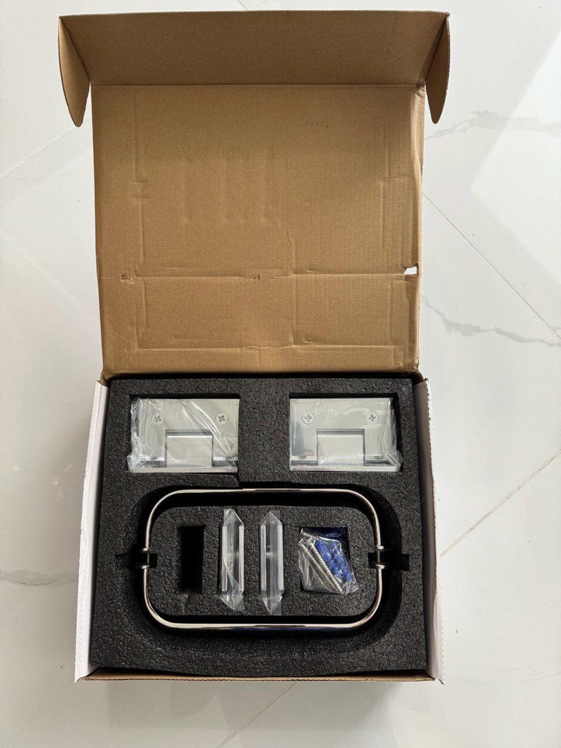 May include: An open cardboard box containing shower door hardware. The set includes two square, clear-wrapped hinges, a curved metal handle, and several small, clear, and blue-wrapped components, all nestled in a black foam insert.