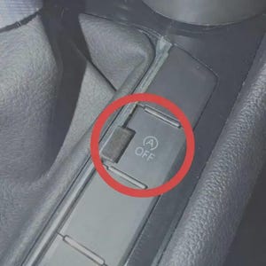 May include: A black plastic car interior button featuring a white icon of the letter A inside a circular arrow with the word OFF printed below it. The button is rectangular and set within a textured black console panel near a gear shift boot.
