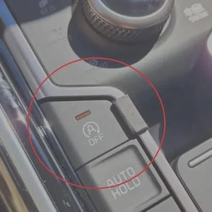 May include: A dark gray vehicle center console button panel featuring an Auto Stop-Start off button with a circular arrow icon and the text OFF, accompanied by a small orange indicator light and a rectangular black plastic cover piece.