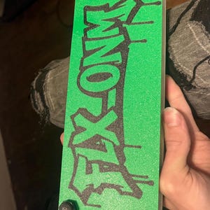 May include: A rectangular green object with the word "FLEX ZONE" in black graffiti-style lettering. The object has four black, rounded corner pieces. The object is being held by a person.