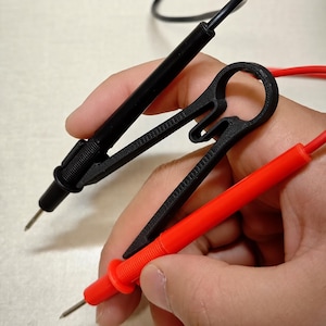 May include: A black 3D-printed plastic multimeter probe holder designed to secure two test leads simultaneously. The holder features a ring-shaped top and two parallel slots that accommodate a black probe and a red probe, each with a metal tip.