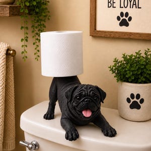 May include: A black pug figurine toilet paper holder with a white roll of toilet paper balanced on its back. The pug has a wrinkled face, dark eyes, and a pink tongue sticking out. The figurine is positioned on a white toilet tank lid.