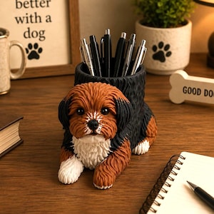 Könnte beinhalten: Ein Schreibtisch-Organizer in Form eines liegenden Hundes mit braunem, schwarzem und weißem Fell. Der Hund hat eine zylindrische Öffnung auf dem Rücken, die mehrere schwarze Stifte hält. Die Figur zeigt detaillierte Fellstruktur und weiße Pfoten.