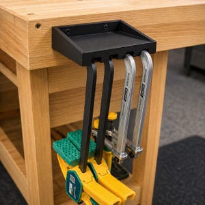 May include: A black metal tool holder mounted on a wooden workbench. The holder has slots for clamps and other tools. Several clamps are hanging from the holder, including yellow and green clamps and silver clamps. The workbench is made of light-colored wood.