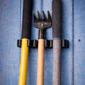 Op de afbeelding: Drie tuingereedschappen, waaronder een hark en twee handgereedschappen, worden getoond tegen een blauwe houten achtergrond. De gereedschappen worden in een zwarte houder gehouden. Eén gereedschap heeft een gele steel, een ander een houten steel en de derde een grijze steel.