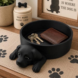 May include: A matte black circular tray featuring a sculpted black puppy head and paws extending from the front edge. Inside the tray are a set of silver metal keys and a small brown leather wallet.