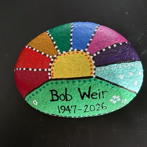 May include: Hand-painted rock with a colorful sunburst design. The sunburst features segments of red, orange, gold, green, blue, purple, and pink, with white dotted accents. The text "Bob Weir 1947-2026" is painted on a green base.